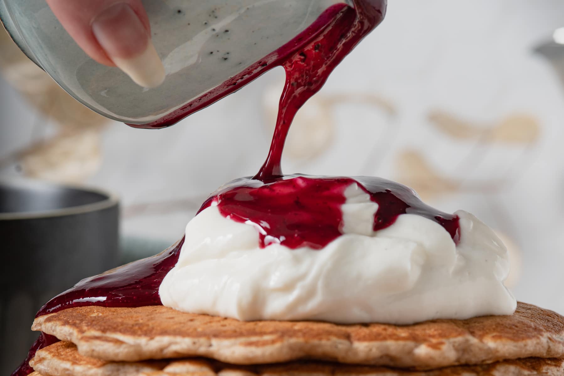 A blueberry syrup being poured from a small jug over pancakes topped with vanilla yogurt.