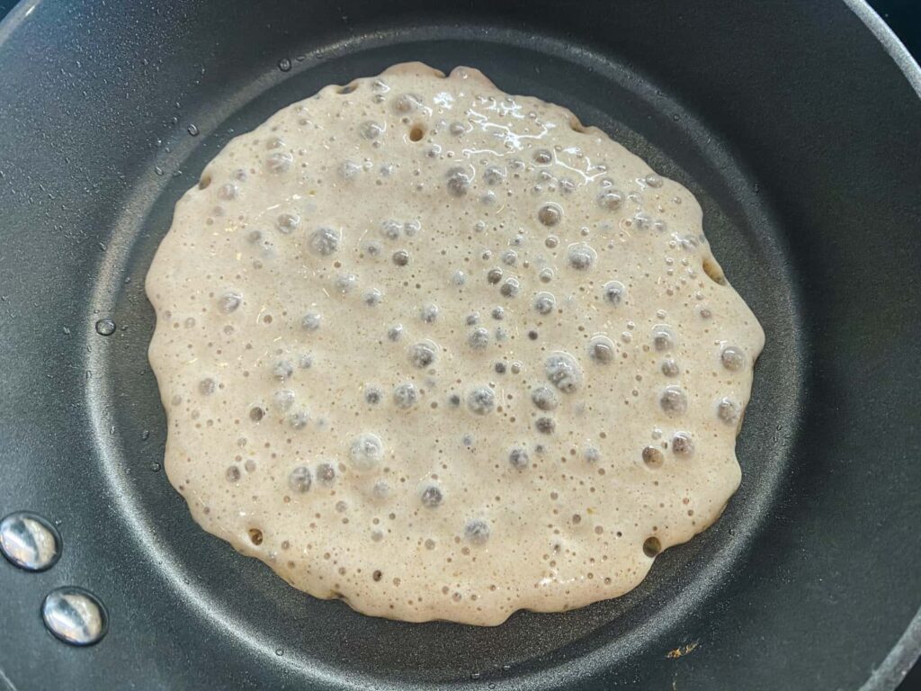 A large buttermilk pancake cooking with lots of air bubbles on the top about ready to be flipped.
