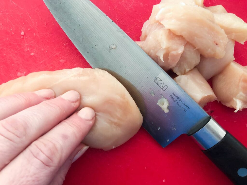 Cutting chicken breasts into small chunks with a sharp knife on a red chopping board.