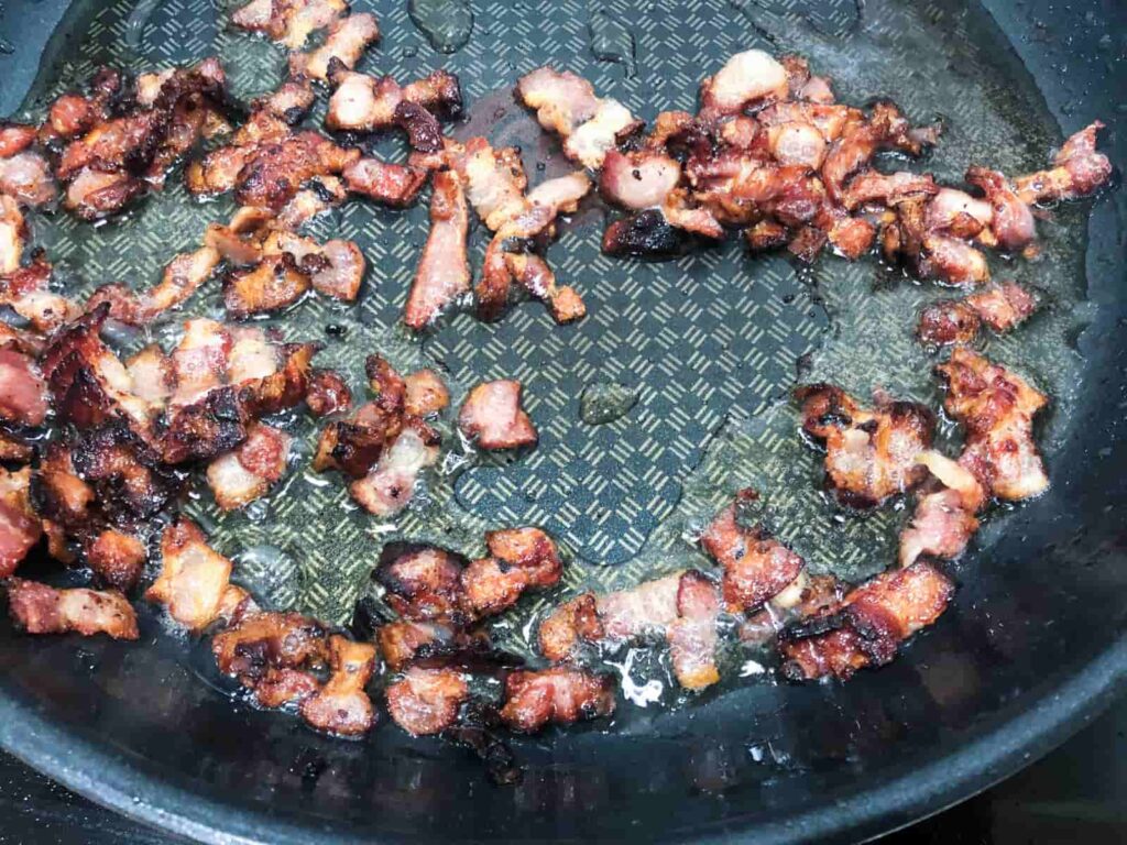 Pan fried pieces of smokey bacon with slight charring around the edges in a large frying pan.