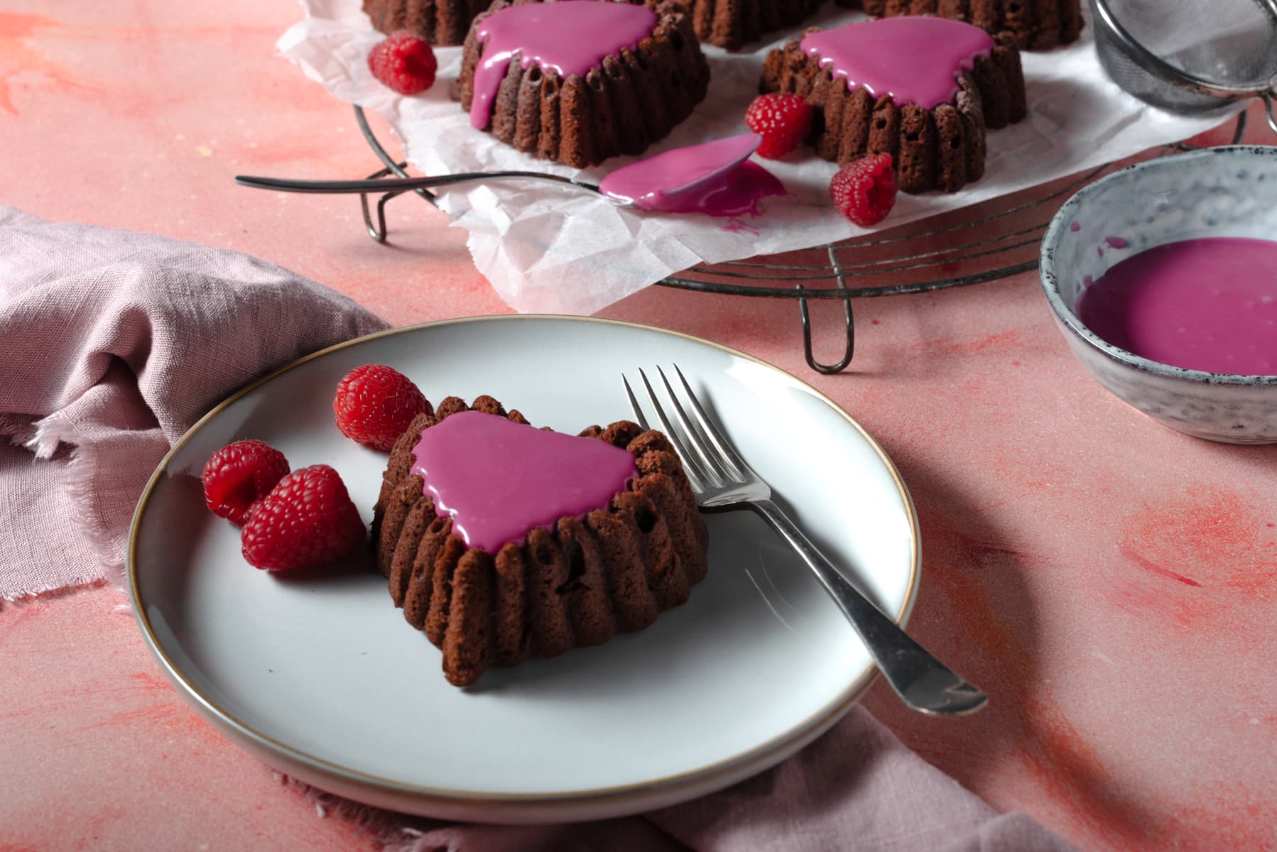 Small chocolate heart shaped cakelettes filled topped with pink icing served with raspberries.