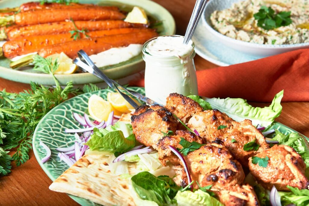 Mezze platter ideas with a plate of marinated chicken skewers, served with a jar of yogurt and lemon sauce, a plate with oven roasted carrots and a bowl of baba ganoush. 