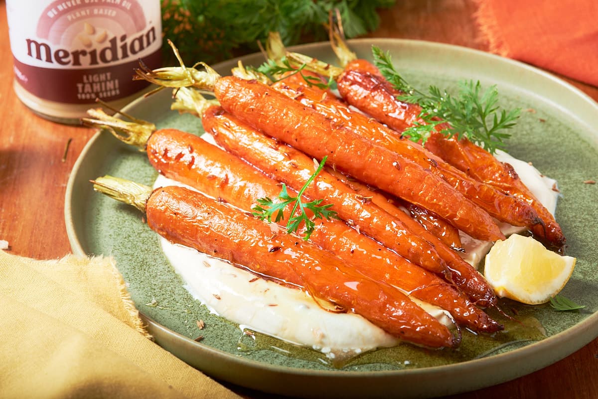 Fresh roasted carrots with honey and cumin seeds, laying on a yogurt tahini dip and sitting on a green plate with orange and mustard coloured napkins around it.