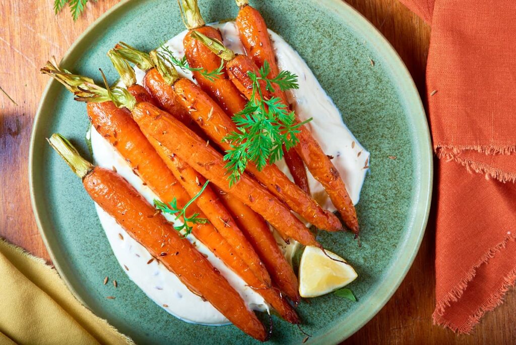 A flatlay of a large green plate topped with roasted carrots laid on a bed of yogurt and lemon sauce with a wedge of lemon.