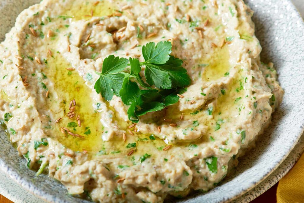 A large grey bowl filled with baba ganoush aubergine dip, topped with a drizzle of olive oil and garnished with fresh parsley.