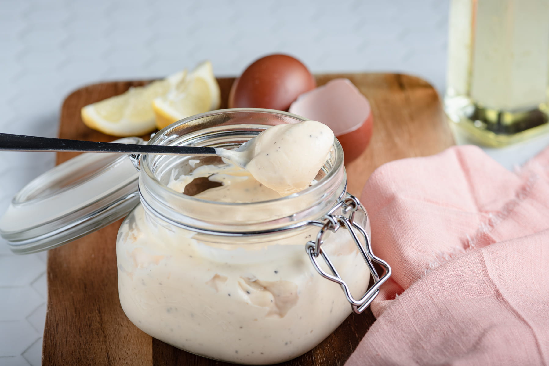 A jar of homemade mayonnaise with lemon slices and eggs to the back on a wooden board in a Kilner flip lid jar.