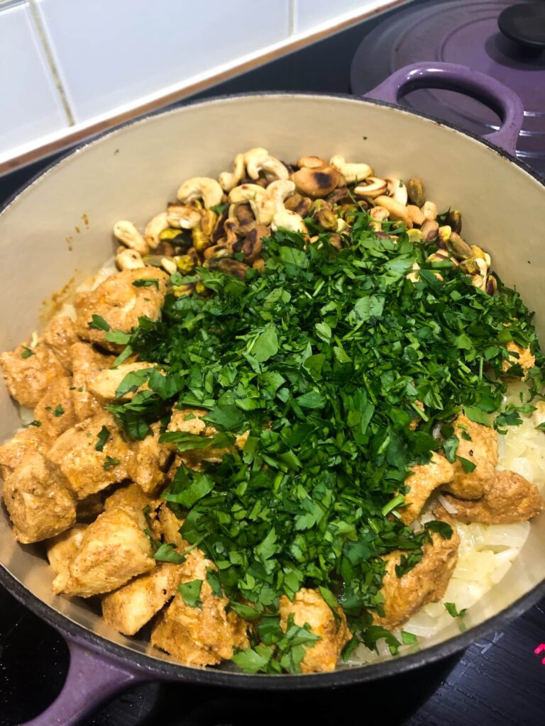 Dutch oven with cooked rice, topped with cooked chicken, toasted nuts and fresh parsley