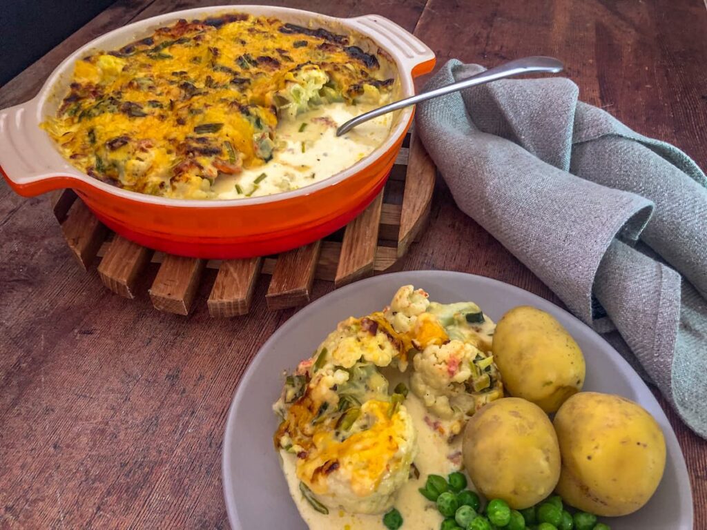 orange oven proof dish filled with gluten free cauliflower cheese on a wooden board and a plate with a serving of cauliflower cheese, with potatoes and garden peas