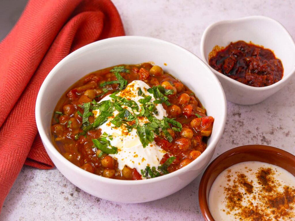 white bowl filled with Moroccan chickpea stew topped with a spoon of Greek yogurt, freshly chopped mind and a drizzle of olive oil with bowls of harissa paste and spiced yogurt set alongside