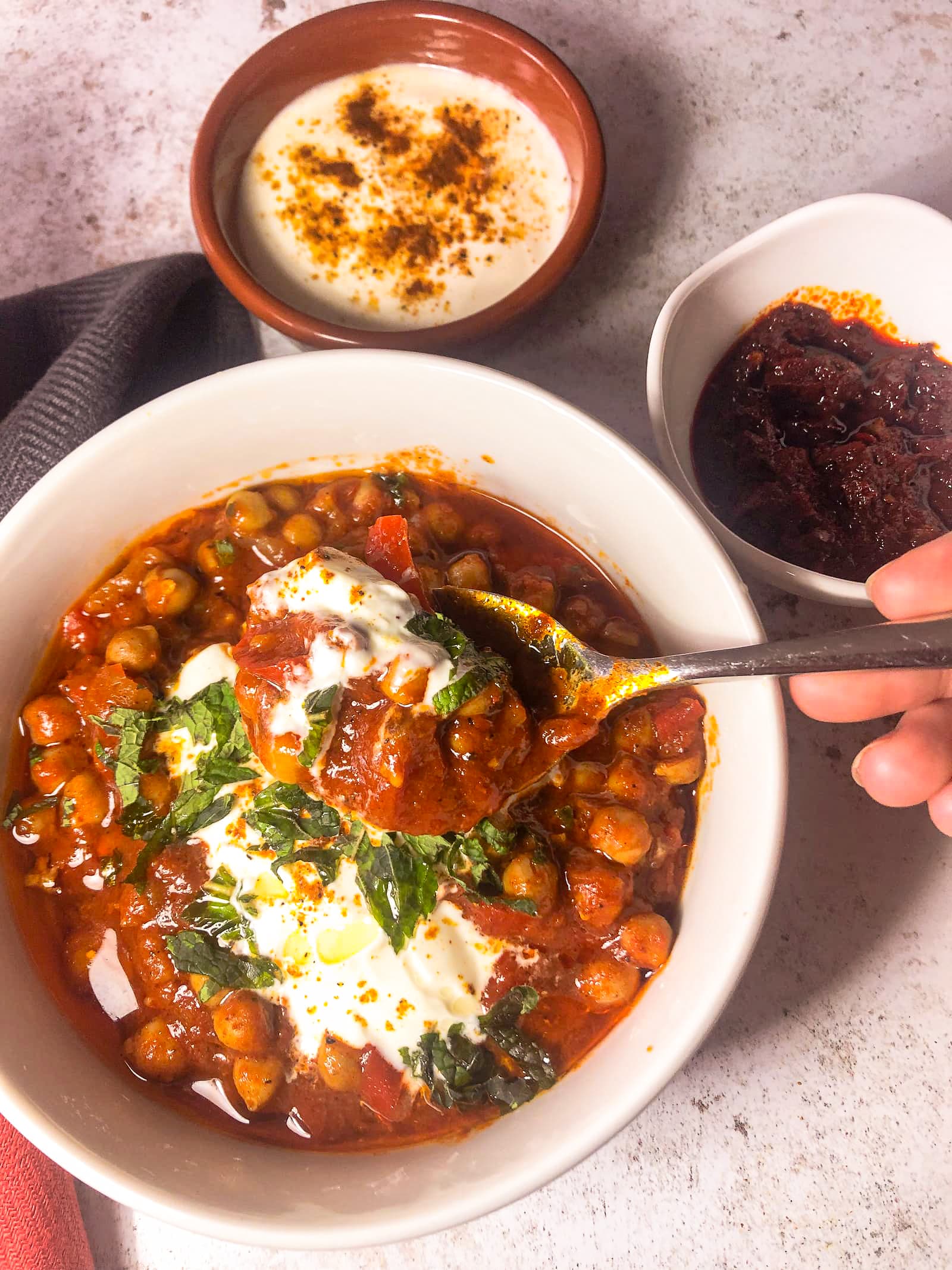 white bowl and spoon filled with Moroccan chickpea stew topped with a spoon of Greek yogurt, freshly chopped mind and a drizzle of olive oil
