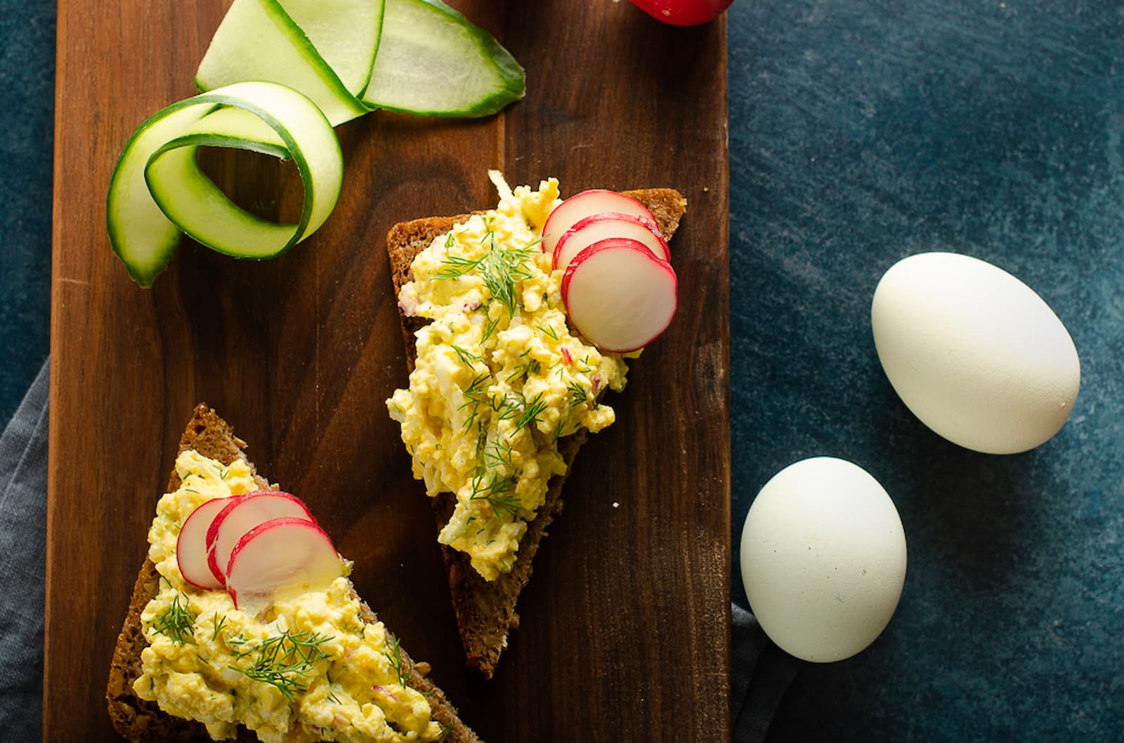wooden board topped with triangles of whole wheat bread topped with Scandinavian egg salad and sliced radishes garnished with freshly chopped dill and 2 fresh eggs set alongside
