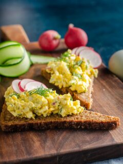 wooden board topped with triangles of whole wheat bread topped with Scandinavian egg salad and sliced radishes garnished with freshly chopped dill