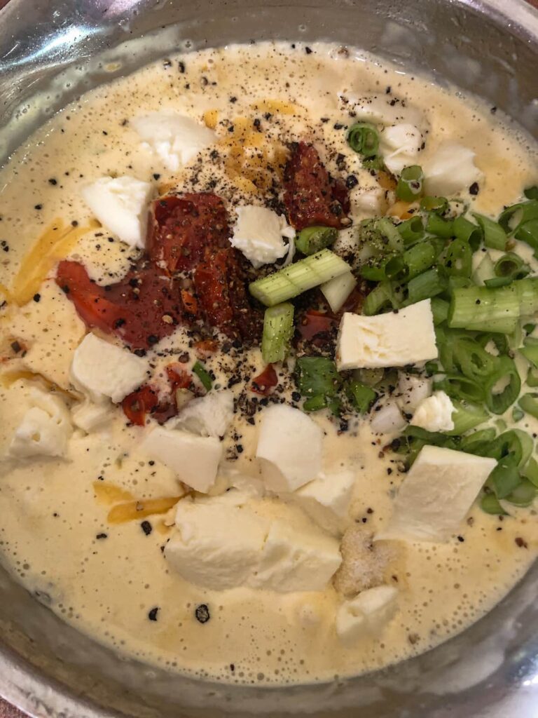 saucepan with creamy cheese sauce topped with sliced spring onions, sun dried tomatoes, cubes of mozzarella cheese and black pepper