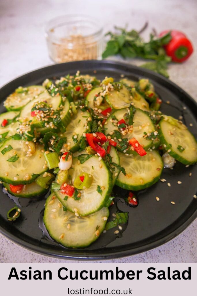 Pinnable image with recipe title and a black plate with sliced cucumbers and topped with diced red chilli, sliced spring onions and toasted sesame seed and coated in salad dressing, with a glass dish filled with toasted sesame seeds, sprigs of fresh herbs and a whole red chilli set alongside.