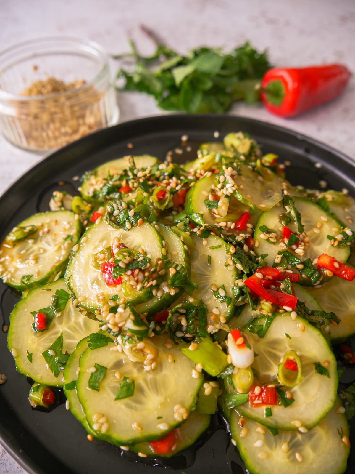 Close up photo of a black plate with sliced cucumbers and topped with diced red chilli, sliced spring onions and toasted sesame seed and coated in salad dressing, with a glass dish filled with toasted sesame seeds, sprigs of fresh herbs and a whole red chilli set alongside.