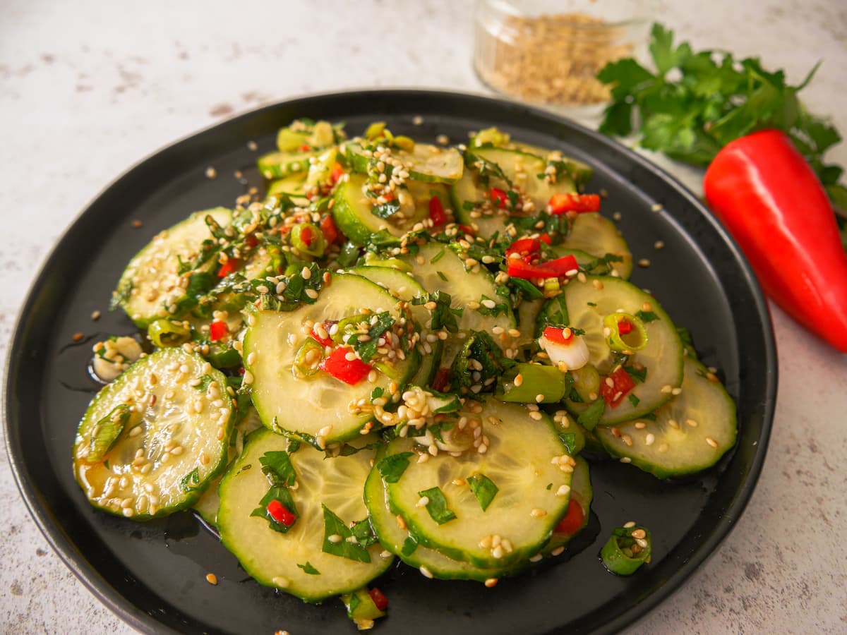 A black plate with sliced cucumbers and topped with diced red chilli, sliced spring onions and toasted sesame seed and coated in salad dressing, with a glass dish filled with toasted sesame seeds, sprigs of fresh herbs and a whole red chilli set alongside.