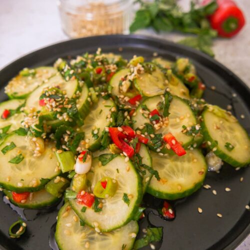 A black plate with sliced cucumbers and topped with diced red chilli, sliced spring onions and toasted sesame seed and coated in salad dressing, with a glass dish filled with toasted sesame seeds, sprigs of fresh herbs and a whole red chilli set alongside.