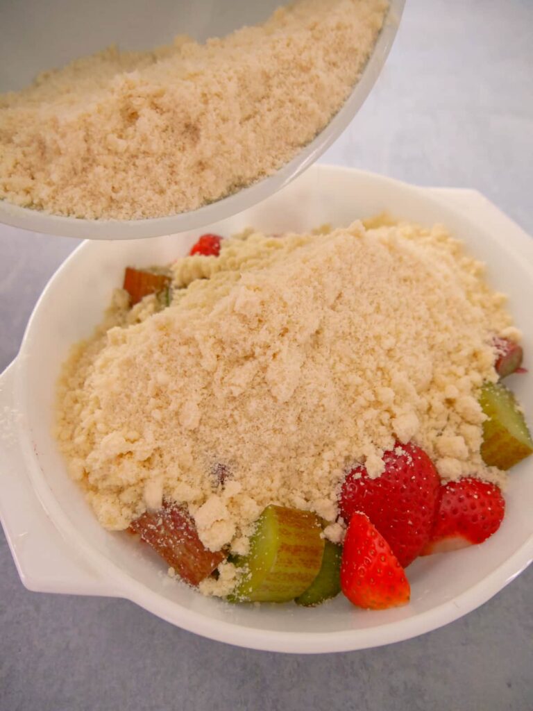 White pyrex bowl with chopped rhubarb and strawberries being covered with crumble topping.