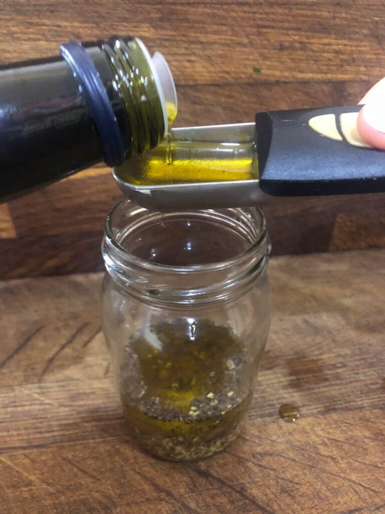 A glass jam jar filled with wholegrain mustard and a measuring spoon being filled with extra virgin olive.