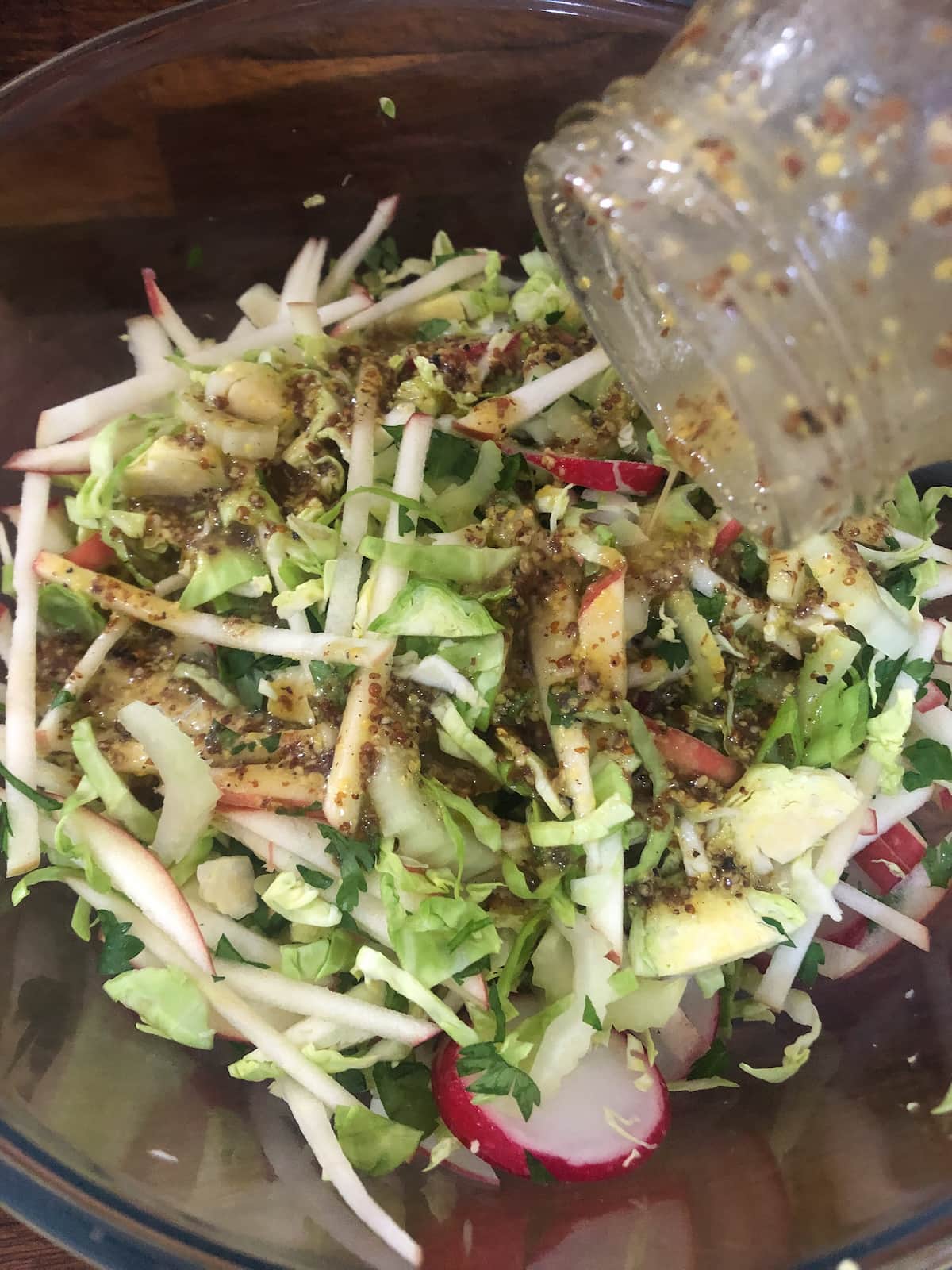A glass bowl filled with shredded sprout slaw ingredients with a jar of honey and mustard salad dressing being poured over.
