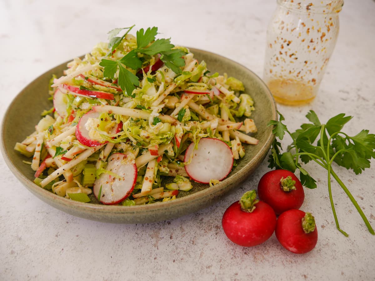 A green bowl filled with dressed shredded Brussels sprouts, red apple, celery and sliced radish, garnished with parsley and an empty jar of dressing, whole radishes and a sprig of parsley set alongside.