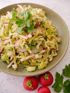 A green bowl filled with dressed shredded Brussels sprouts, red apple, celery and sliced radish, garnished with parsley and whole radishes and a sprig of parsley set alongside.