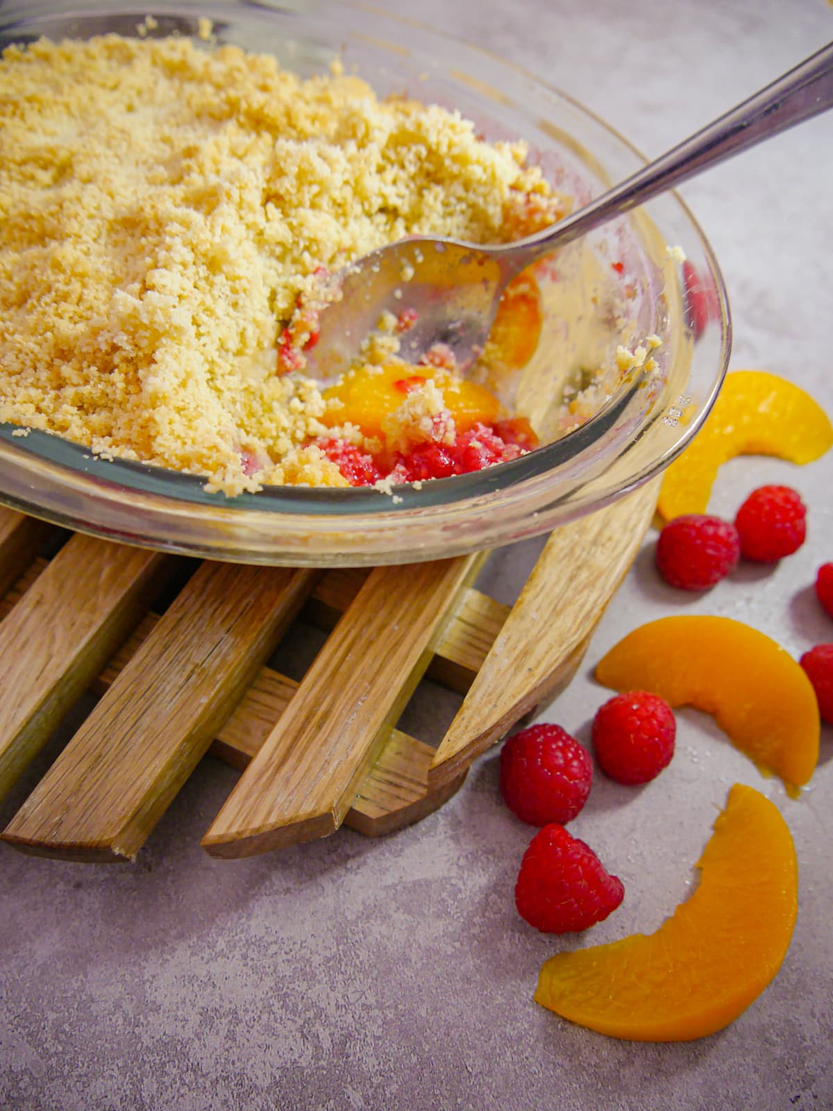 close up of peach and raspberry crumble set on a wooden board with a serving spoon and portion removed from dish and fresh raspberries and peach slices alongside