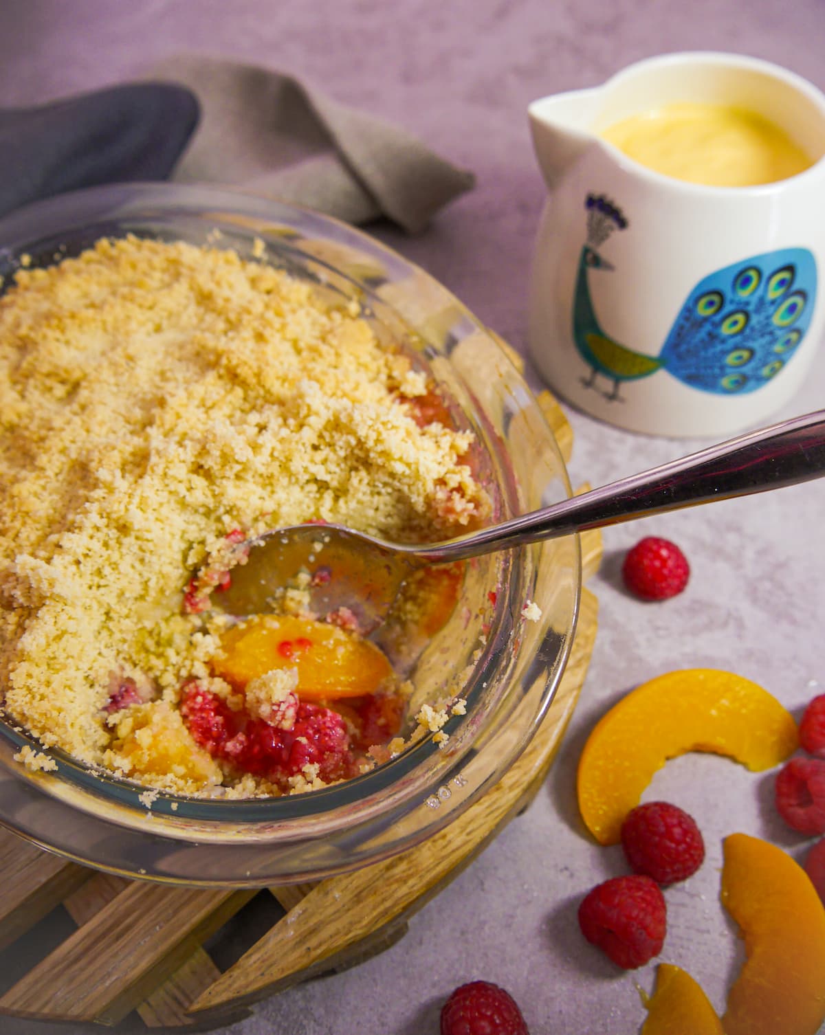 close up of peach and raspberry crumble set on a wooden board with a serving spoon and portion removed from dish, with a jug of custard and fresh raspberries and peach slices set alongside
