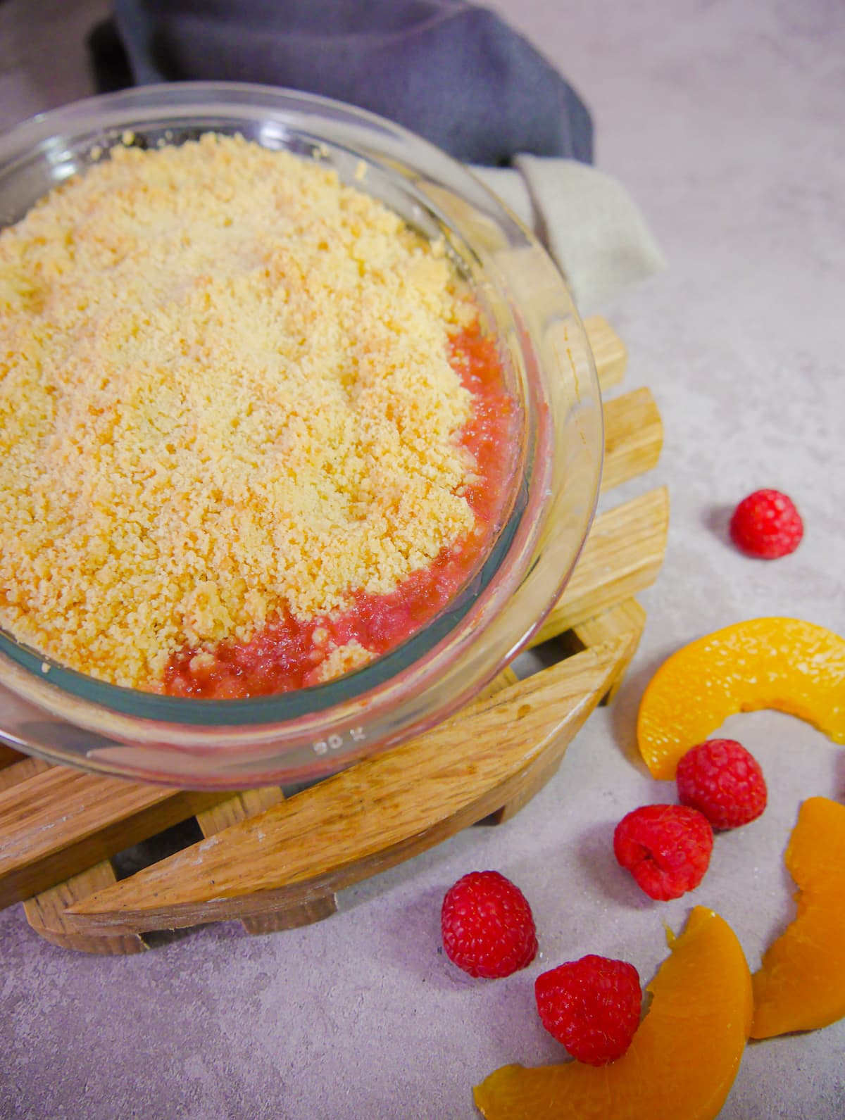 glass dish of peach and raspberry crumble set onto a wooden trivet with peach slices and fresh raspberries set alongside