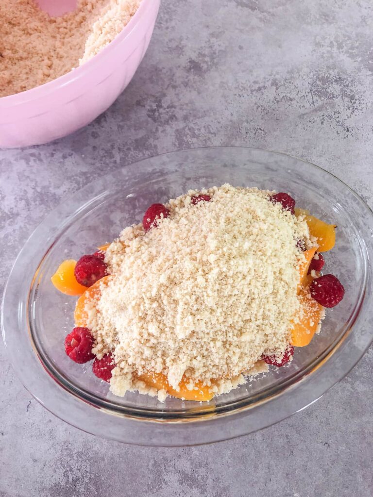 glass bowl filled with sliced peaches and fresh raspberries topped with crumble topping