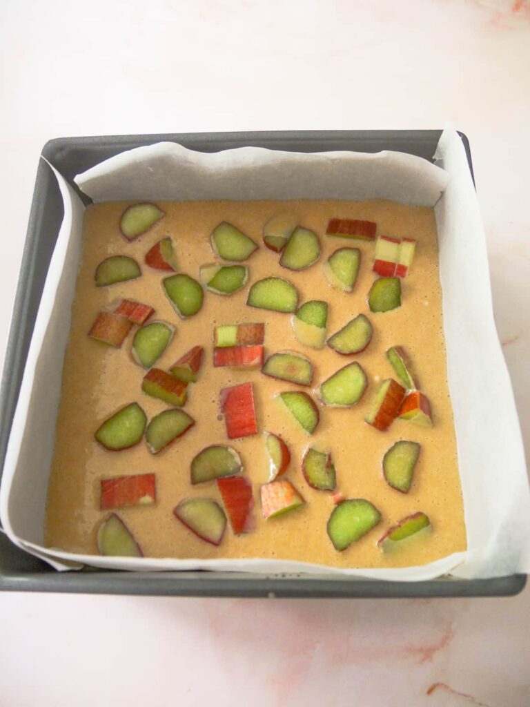 A square baking tin lined with baking paper, filled with rhubarb and ginger cake batter and topped with slices of fresh rhubarb.
