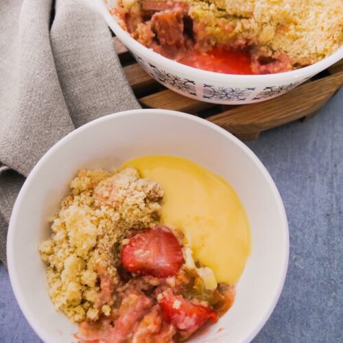 White pyrex dish filled with rhubarb and strawberry crumble, set on a wooden board, with a while bowl of crumble and custard set alongside.