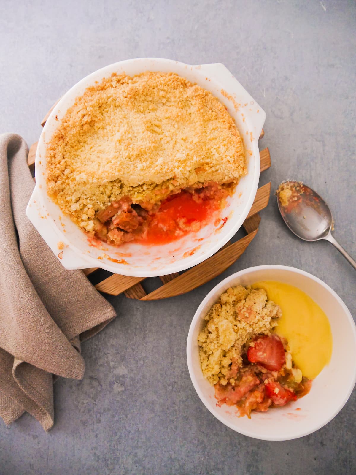 White pyrex dish filled with rhubarb and strawberry crumble, set on a wooden board, with a while bowl of crumble and custard set alongside.