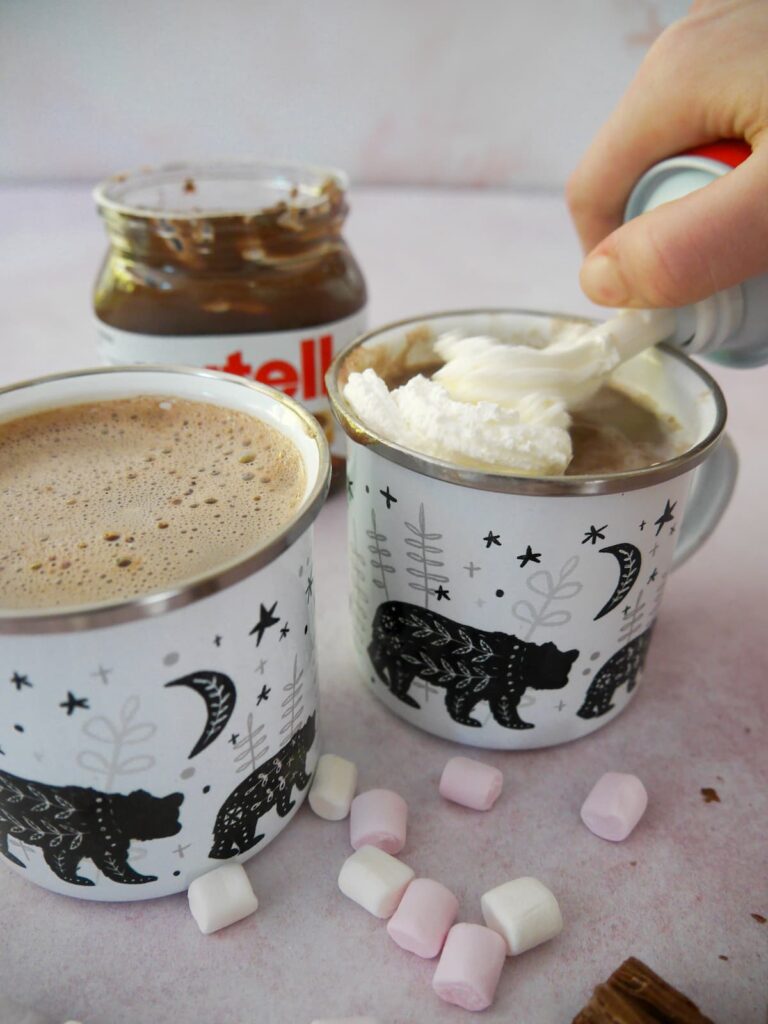 Two mugs of hot chocolate being topped with cream, with an open jar of Nutella, mini marshmallows and chocolate logs set alongside.