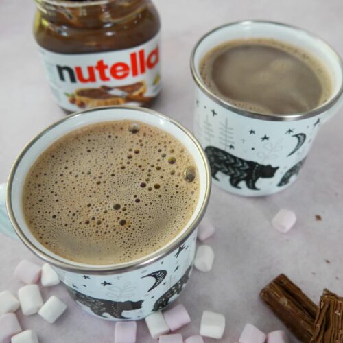 Two mugs of hot chocolate with an open jar of Nutella, mini marshmallows and chocolate logs set alongside.