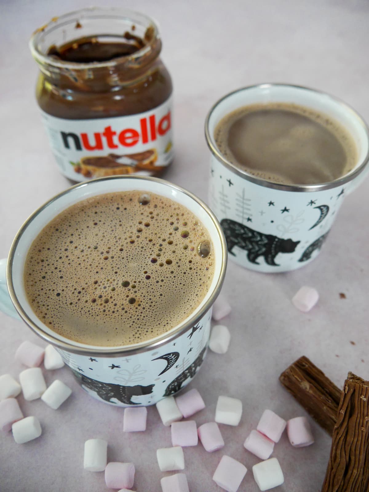Two mugs of hot chocolate with an open jar of Nutella, mini marshmallows and chocolate logs set alongside.