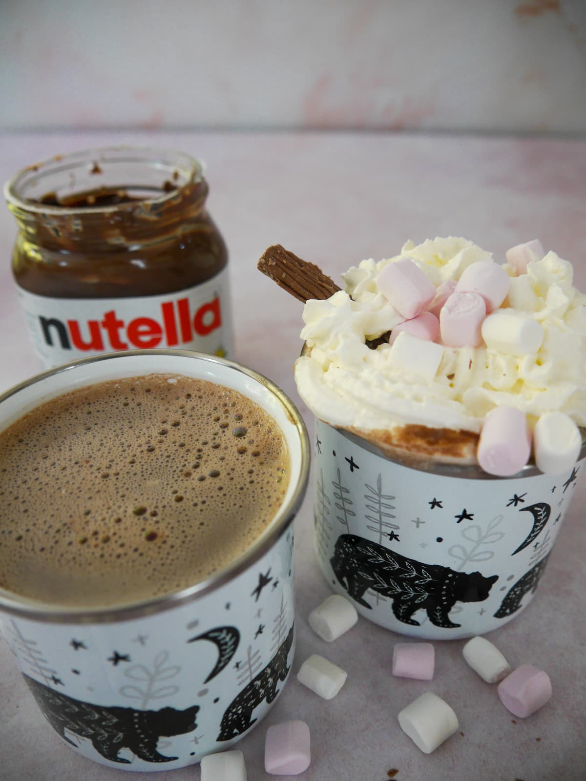 Two mugs of hot chocolate, one topped with whipped cream, mini marshmallows and a chocolate log, with an open jar of Nutella, mini marshmallows and chocolate logs set alongside.