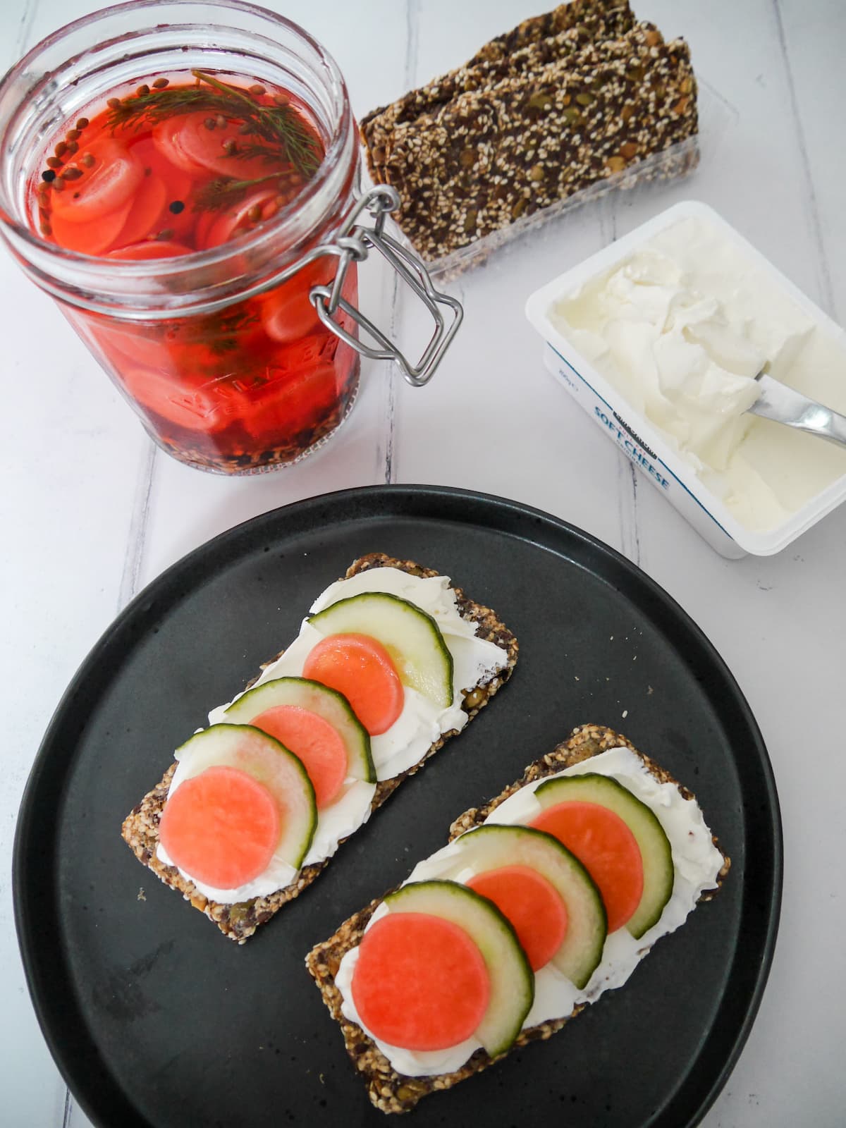 A black plate with seeded crackers, topped with cream cheese and slices of cucumber and pickled radish; and a jar of radishes, a box of crackers and a tub of cream cheese set alongside.