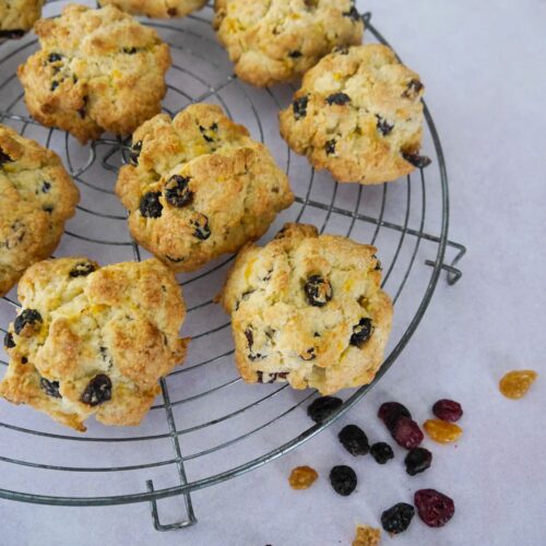 A circular wire rack topped with rock cakes, and some dried raisins, golden raisins and cranberries scattered alongside.