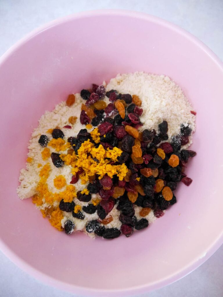 A pink bowl filled with crumbed rock cake base topped with grated orange zest and a mixture of dried fruits.