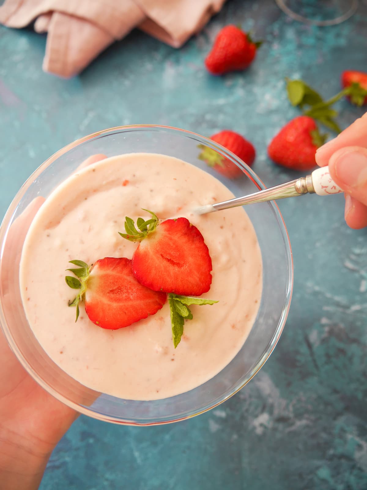 A hand holding a glass serving dishes filled with strawberry mousse topped with sliced strawberries and mint leaves, with whole strawberries set alongside.