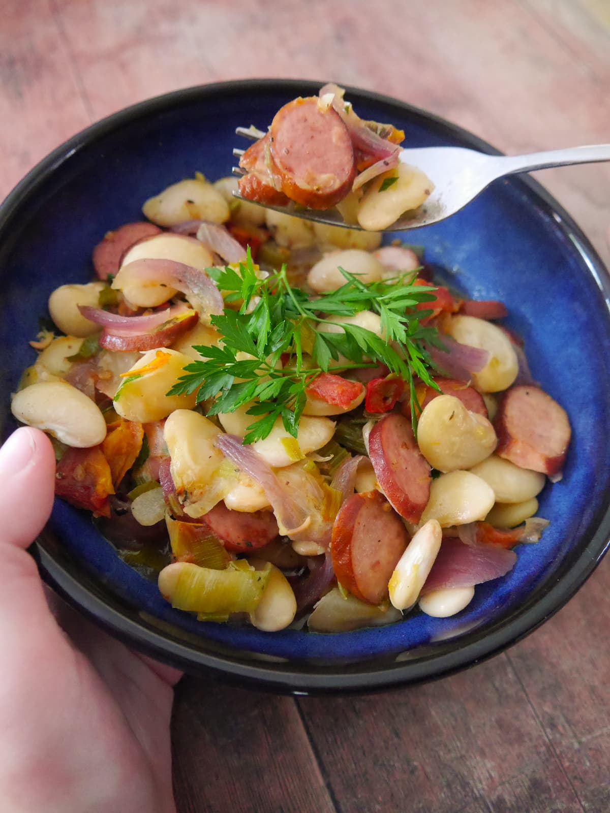 Hands holding a fork and blue bowl filled with butter bean and smoked sausage stew with a garnish of fresh parsley.