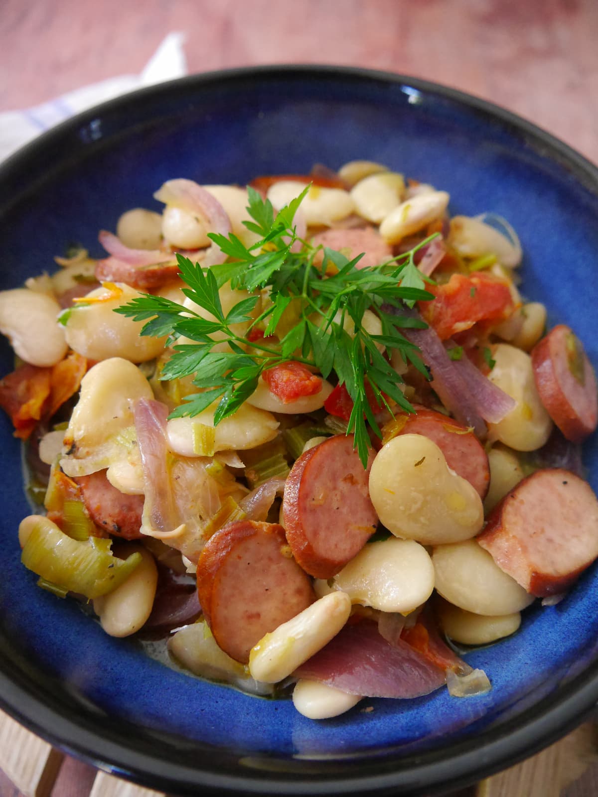 Close up image of blue bowl filled with butter bean and smoked sausage stew with a garnish of fresh parsley.