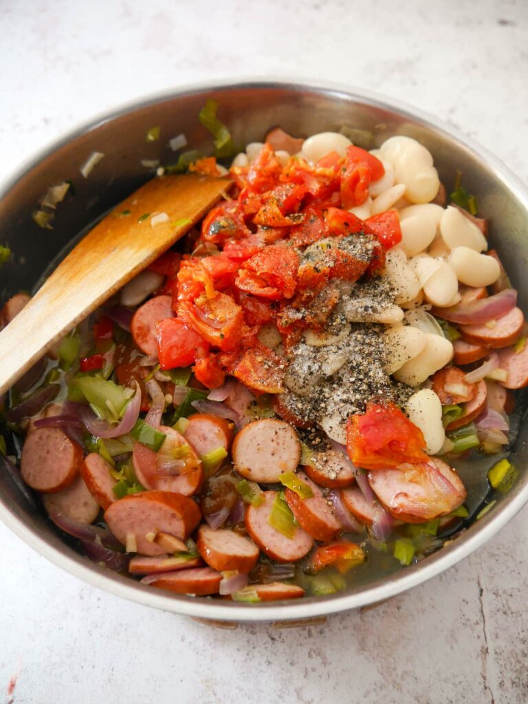 A frying pan of sauteed vegetables and smoked sausage topped with chopped sun blush tomatoes and canned butter beans.