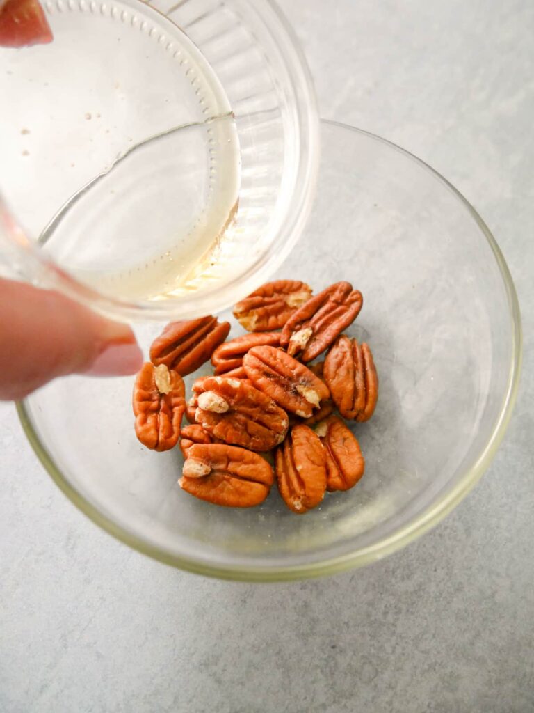 Glass bowl filled with candied pecans with maple syrup being poured over.