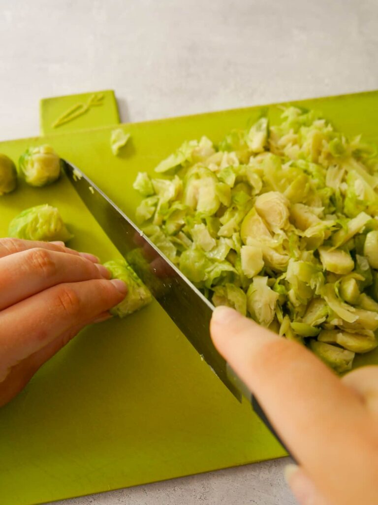 A pair of hands holding a kitchen knife chopping up Brussels sprouts on a board.
