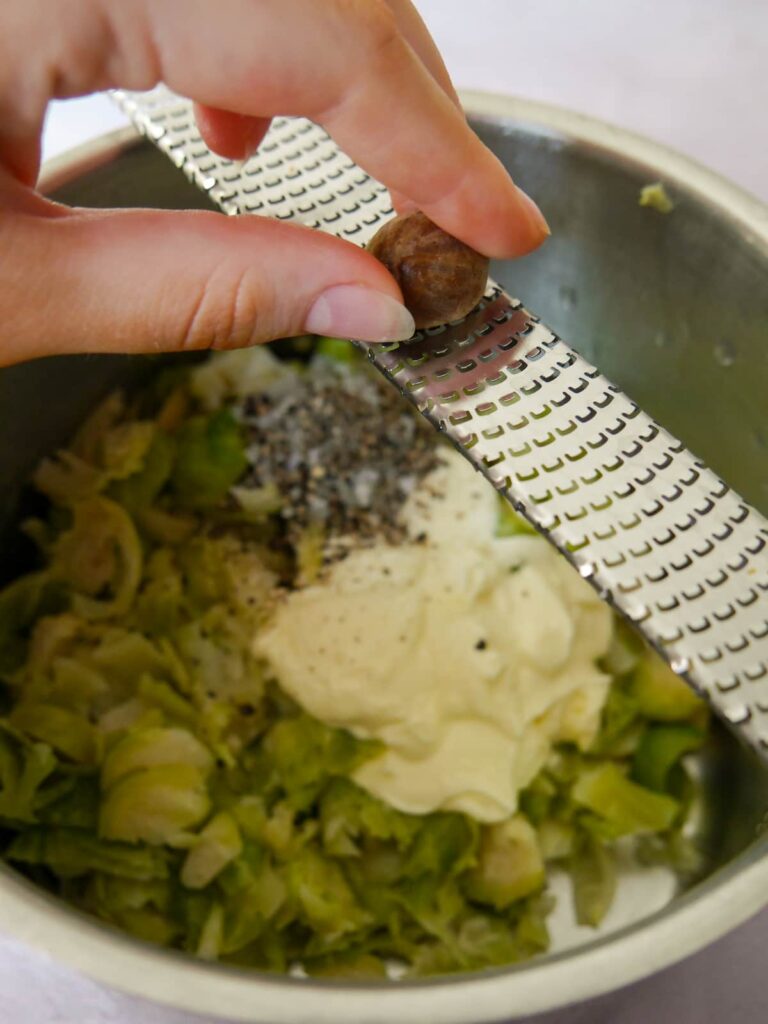 A hand holding a grater and whole nutmeg being grated over a saucepan of shredded Brussels sprouts.