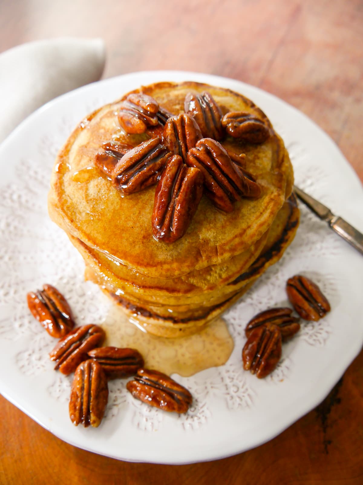 A white plate with a stack of pumpkin pancakes topped with candied pecans and a drizzle of maple syrup.