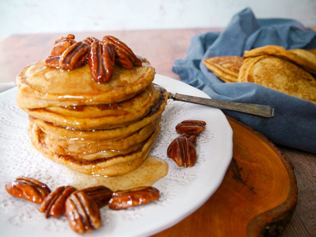 A stack of pumpkin pancakes on a white plate, topped with candied pecans and maple syrup, with a plate of pumpkin pancakes set alongside.
