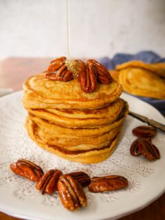 A white plate with a stack of pumpkin pancakes topped with candied pecans and a drizzle of maple syrup being poured over.
